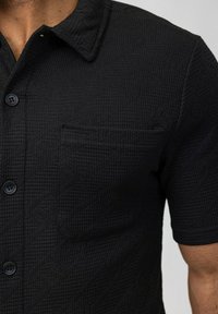 Chemise noire à manches courtes en tissu texturé avec un motif subtil, dotée d'un col boutonné et d'une poche poitrine.