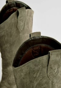Botas de vaquero de ante en verde oliva con patrones de hojas embossados. Forro marrón y detalles de costura. Incluye lengüetas para facilitar el uso.