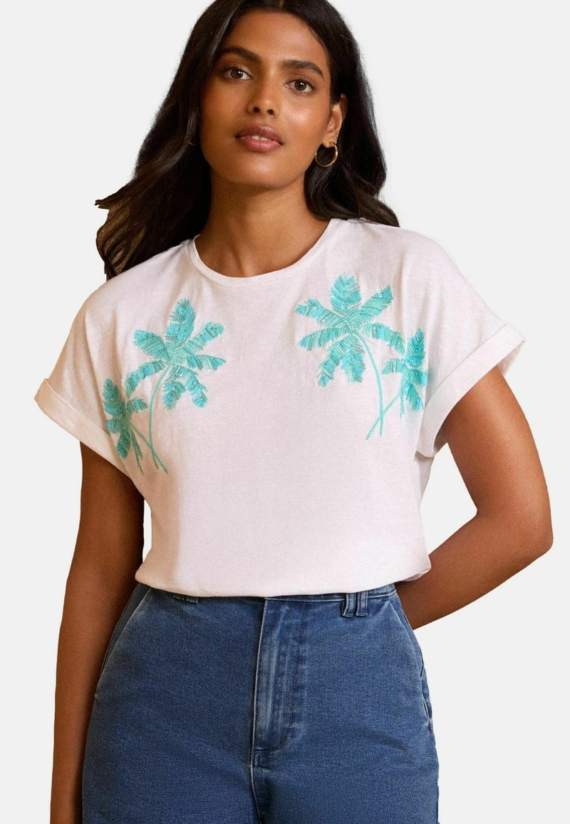 Baltas kokvilnas T-krekls ar sarullētām īsām piedurknēm, uz kura priekšpuses ir turkoza izšūtas palmas. Apvienots ar augsta vidukļa zilajiem džinsiem.