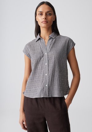 Femme aux longs cheveux foncés portant une chemise à manches courtes grise à motifs et un pantalon foncé, debout devant un fond uni.