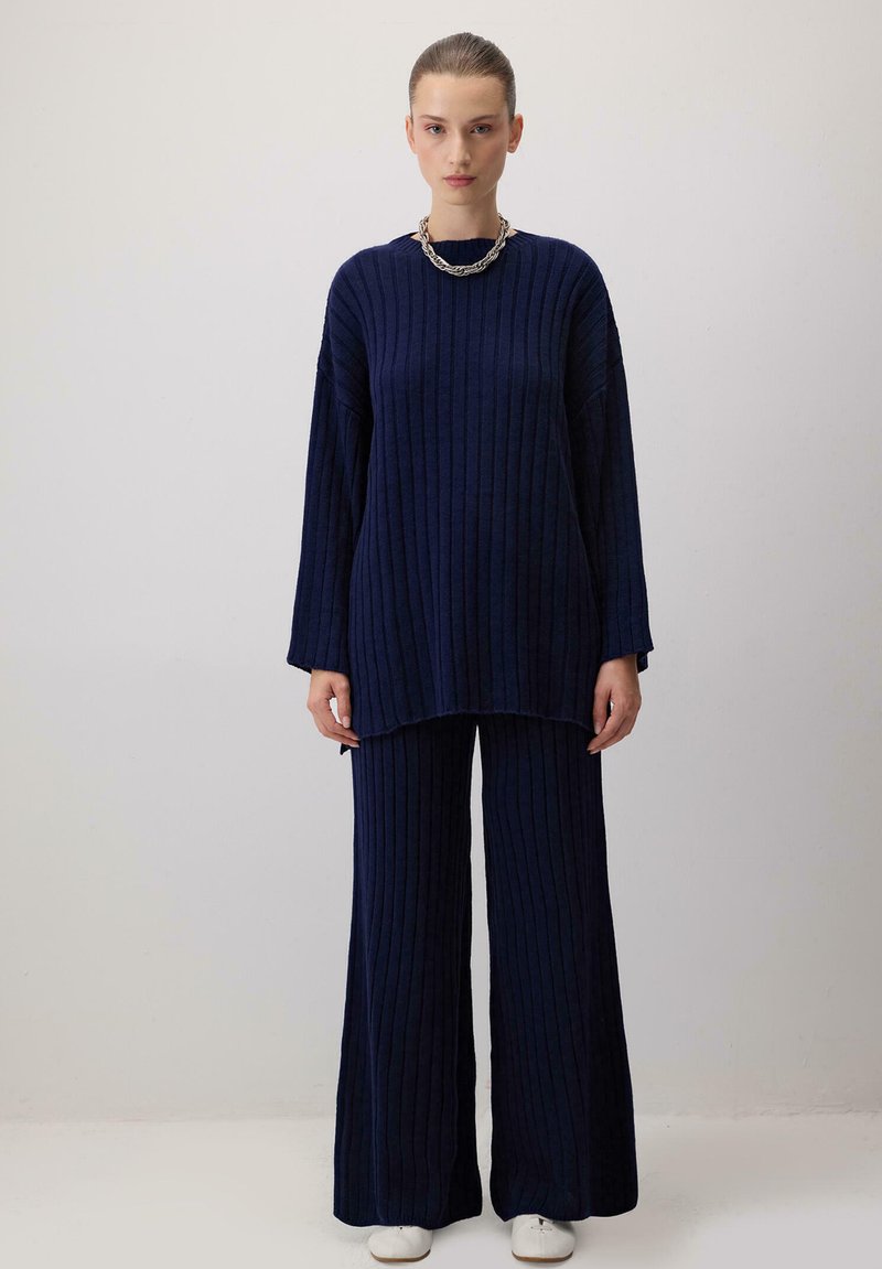 Tumes ribakootud sviiter langevate õlgadega ja ümmarguse kaelusega, millele on lisatud laiad püksid; mõlemad on lõdva lõikega ja pehme tekstuuriga.