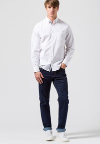 Camisa blanca de botones con un bolsillo delantero, combinada con jeans azul oscuro y zapatillas blancas. La camisa tiene mangas largas y un cuello clásico.