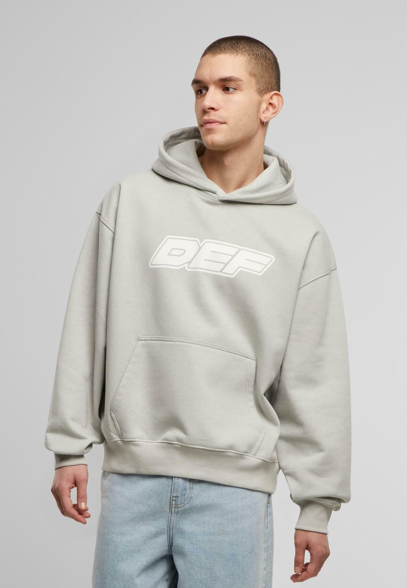 Lys grå hættejakke med en frontlomme, der har et stort hvidt "DEF" logo. Designet har en afslappet pasform og blød fleecetekstur.