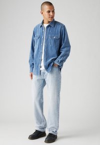 Lichtblauwe corduroy-shirt met knoopsluiting en twee borstzakken, gecombineerd met lichtblauwe jeans en zwarte schoenen, staande tegen een neutrale achtergrond.