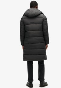 Abrigo acolchado negro con capucha, presenta un diseño acolchado y un logo en la parte posterior. Combina con pantalones oscuros y zapatos negros.