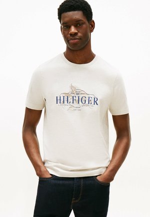 Lys beige bomulds t-shirt med et navyblåt og gyldent trykt logo. Rund hals og korte ærmer, parret med mørk denim jeans.