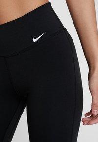 Leggings atletici neri realizzati in tessuto elasticizzato con vita alta. Presentano un logo Nike bianco sulla parte anteriore e dettagli di cucitura visibili.