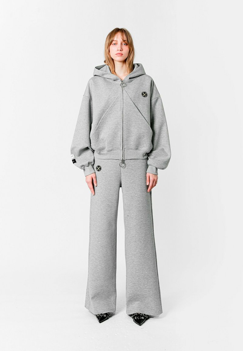 Sudadera gris claro, de gran tamaño, con cremallera frontal y detalle con logo, combinada con pantalones de chándal de pierna ancha a juego. Ambos hechos de una tela suave y texturizada.