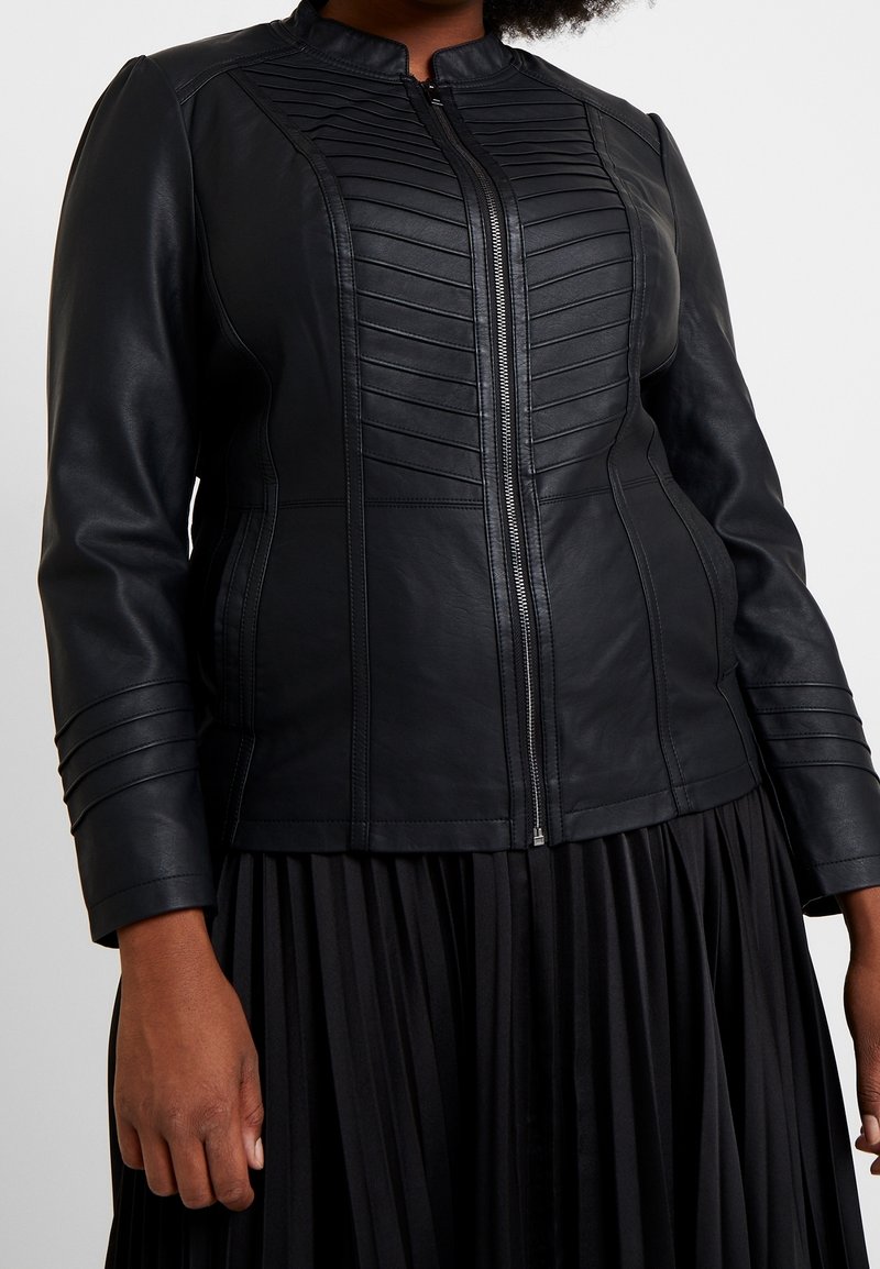 Veste en cuir noire ajustée avec des panneaux cousus horizontalement sur la poitrine et les manches, fermeture éclair frontale, portée avec une jupe plissée noire.