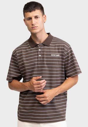 Young man with short hair wearing brown and white striped polo shirt with "never quit" text, standing against a plain light background.