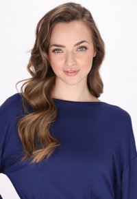 Námořnická modrá volná košile s širokým výstřihem, vyrobená z měkké látky. Modelka má dlouhé vlnité vlasy a je umístěna uprostřed na světlejším pozadí.