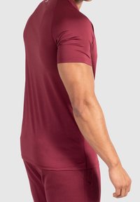 Burgundy idrottströja, figurnära design, korta ärmar, mjuk textur. Har en rundad halsringning och subtil logotypdetalj på baksidan.