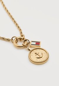 Κολιέ σε χρυσούς τόνους με στρογγυλό μενταγιόν που απεικονίζει άγκυρα και το κείμενο "Tommy Hilfiger", καθώς και ένα μικρό ορθογώνιο γούρι με τη σημαία σε κόκκινο, λευκό και μπλε.