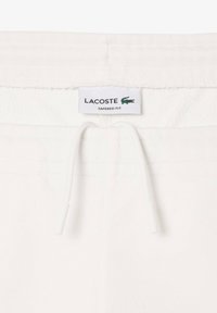 Beli beli slim-fit trenirke v beli barvi z mehkim teksturo, elastičnim pasom in vrvico za zategovanje. Imajo oznako logotipa Lacoste na pasu.