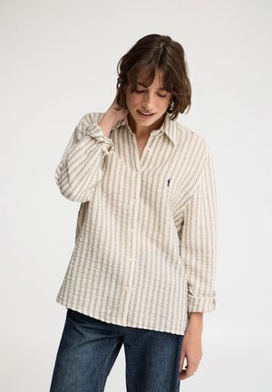 Jeune femme aux cheveux bruns courts portant une chemise beige à rayures avec boutons et un jean foncé, touchant son cou et regardant vers le bas.