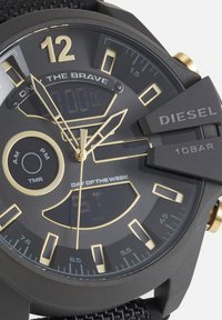 Cadran de montre Diesel noir et or affichant les aiguilles des heures, minutes, secondes, un affichage numérique et un indicateur du jour de la semaine avec un bracelet noir texturé.