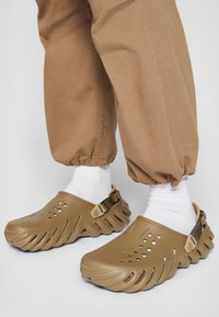 Brown textured clogs feature a curved design with ventilation holes and a back strap. Worn with white socks and tan, elastic cuffed trousers.
