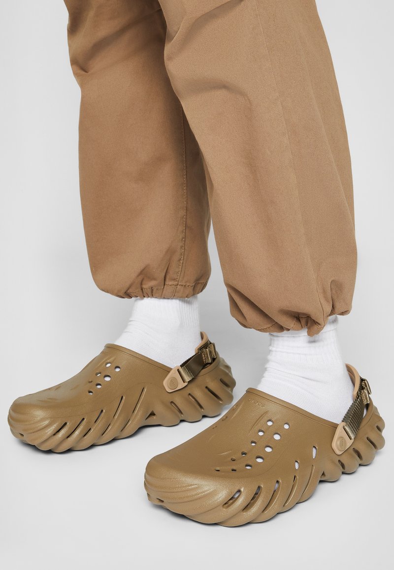 Brown textured clogs feature a curved design with ventilation holes and a back strap. Worn with white socks and tan, elastic cuffed trousers.