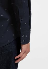 Camicia blu navy con motivo a triangoli bianchi; maniche lunghe, polsini con bottoni e texture liscia. Presenta tre bottoni sul polsino.
