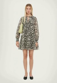 Rochie florală alb-negru, cu mâneci lungi și vaporoase, decolteu rotund și detaliu în formă de cheiță, asortată cu o geantă verde deschis și încălțăminte plată.