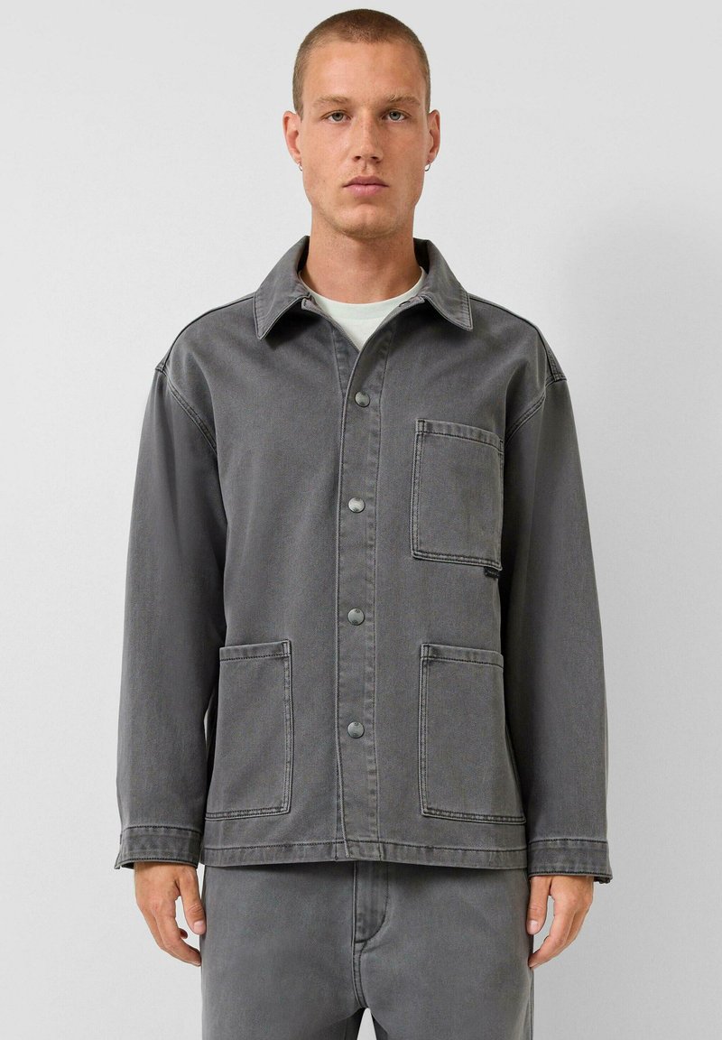 Man draagt een grijze jas met knopen en drie zakken aan de voorkant, gecombineerd met een bijpassende grijze broek, staand tegen een effen lichte achtergrond.