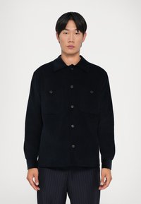 Tumman navy sininen corduroy-paitatakki, jossa on napit etupuolella, kaksi rintataskua ja kaulus. Pitkissä hihoissa on napilliset hihansuut ja teksturoitu pintakuvio.