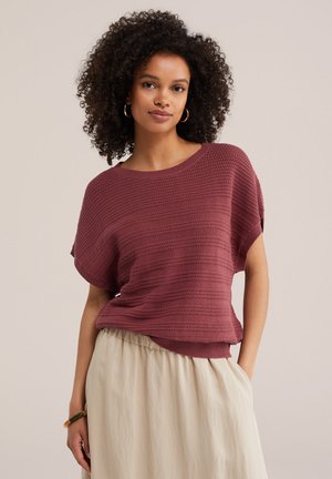 Femme aux cheveux bouclés portant des boucles d'oreilles créoles en or, un pull texturé mauve à manches courtes, une jupe beige et un bracelet à son poignet gauche.