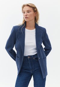 Tmavě modrý, strukturovaný blazer s jedním knoflíkem, klopami a dvěma předními kapsami, spárovaný s bílou košili a modrými džíny.