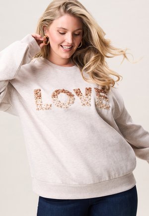 Lachende vrouw die gouden oorring aanraakt, gekleed in een lichtbeige sweatshirt met "LOVE" in parel- en kraalversieringen, gecombineerd met een donkere spijkerbroek.