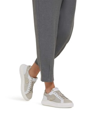 Pies con zapatillas deportivas bajas beige y blancas con suelas blancas, combinadas con pantalones grises ajustados, de pie sobre un fondo blanco.