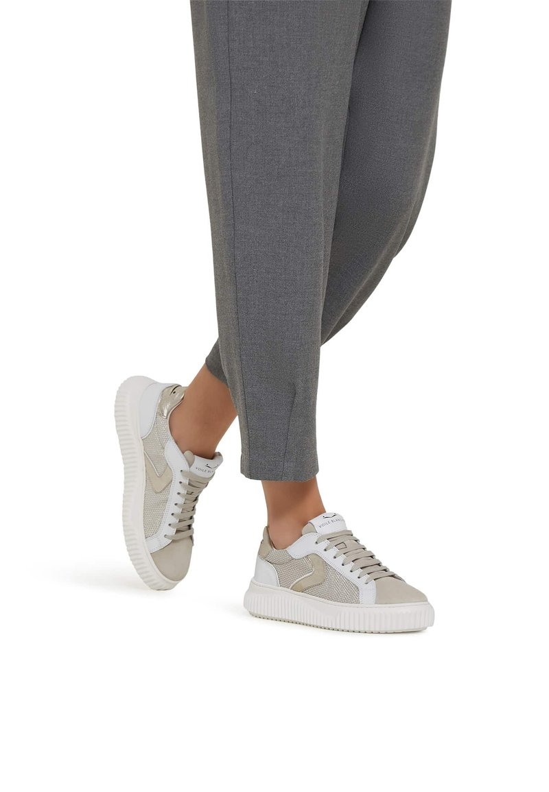 Pieds chaussés de baskets basses beige et blanches avec des semelles blanches, associées à un pantalon gris fuselé, debout sur un fond blanc.