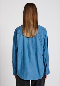 Blå denimskjorta med avslappnad passform, långa ärmar, en veck bak och knappade manschetter. Slät textur, avslappnad design.