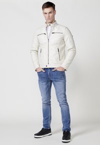 Chaqueta de cuero blanca con bolsillos con cremallera, cuello alto y detalles acolchados, combinada con jeans azules y zapatillas negras.