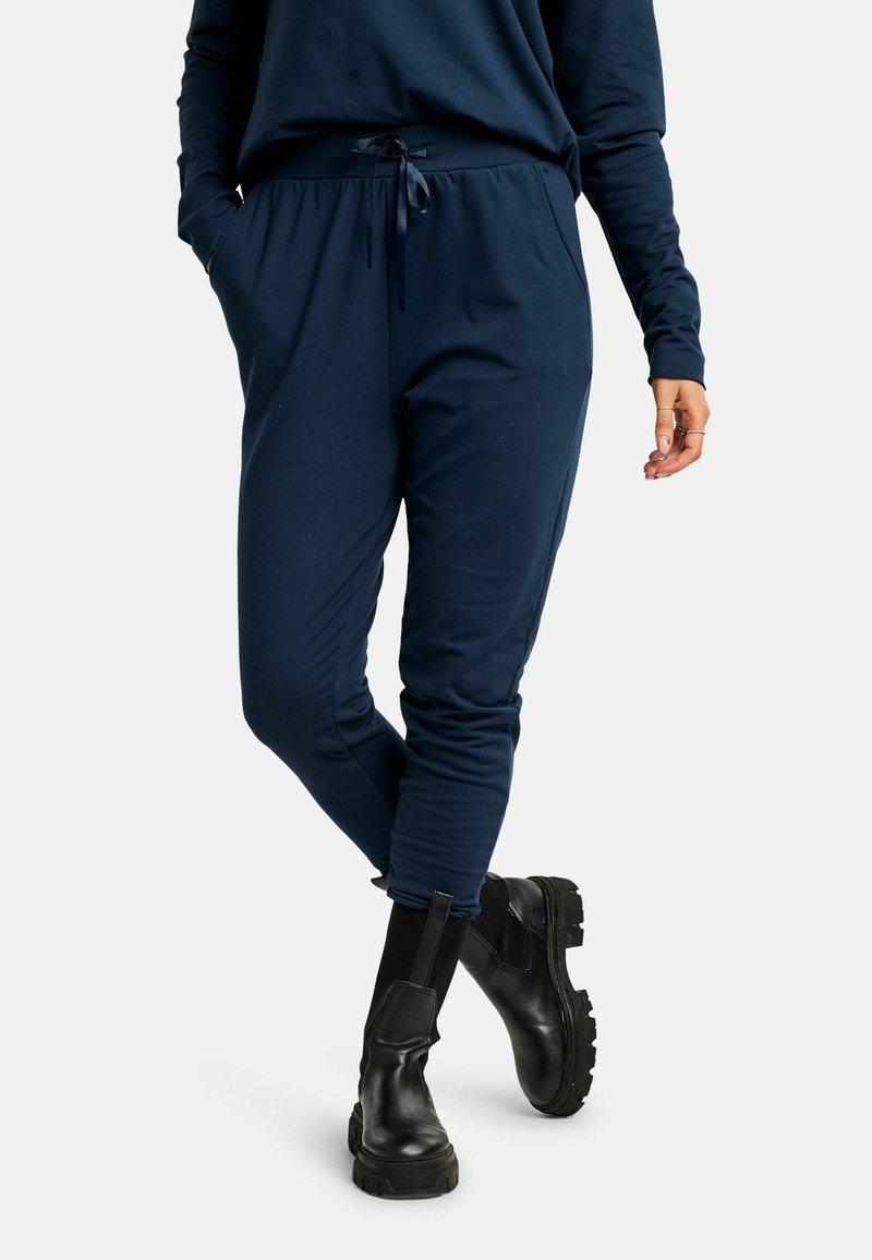 Námornícky modré teplákové nohavice z ľahkej tkaniny, s elastickým pásom so sťahovacou šnúrkou a bočnými vreckami, v kombinácii s čiernymi členkovými čižmami.