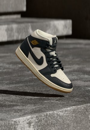Μαύρο και λευκό sneaker Nike Air Jordan με λογότυπο σε χρυσό, ψηλά στον αέρα πάνω από σκάλες από σκυρόδεμα, ρίχνοντας σκιά στο έδαφος από κάτω.