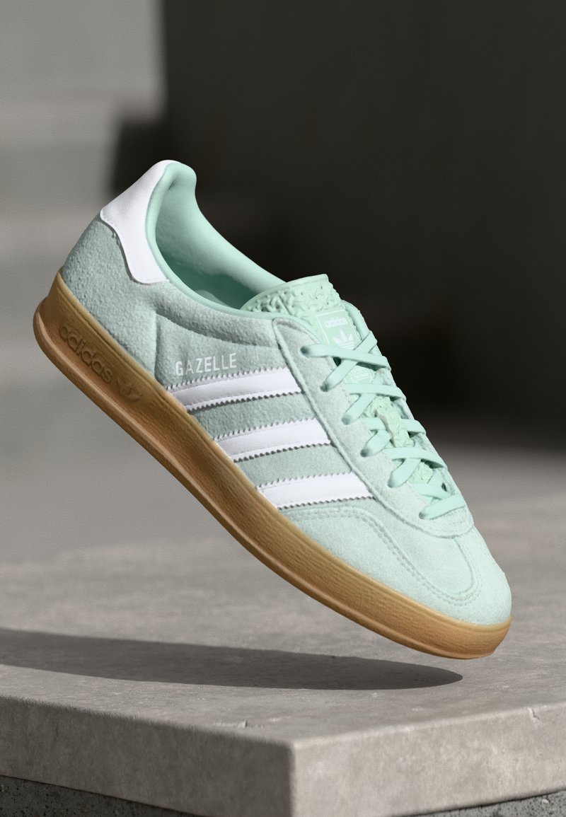 Tenisky Adidas Gazelle v mintově zelené nubukové kůži s bílými akcenty, šněrováním a gumovou podrážkou. Obsahuje tři bílé pruhy.