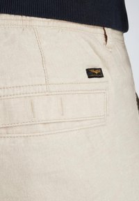 Lichtbeige pantalons met een textuurstof, voorzien van een achterzak en een zwart label met een gouden embleem. Gestikte details zijn zichtbaar.