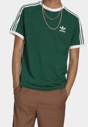 Persoon draagt een groen Adidas T-shirt met witte strepen op de mouwen en logo, bruine broek en gouden kettingen, handen in de zakken.