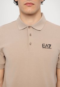 Beige polo skjorte laget av myk stoff, med klassisk krage, tre knapper og logo på venstre bryst. Korte ermer, vanlig passform.
