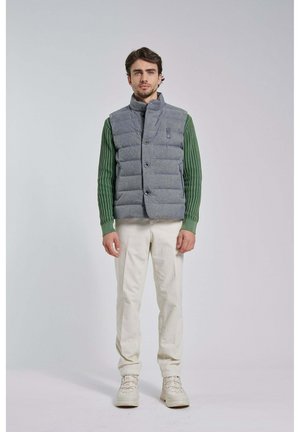 Uomo in piedi con indosso una camicia verde lavorata a maglia a maniche lunghe, un gilet imbottito grigio, pantaloni beige chiaro e stivali beige con lacci su uno sfondo uniforme.