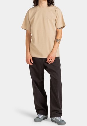 Beige kortærmet t-shirt med rund halsudskæring og logodetaljer, kombineret med sorte bukser med vide ben og grå sneakers.