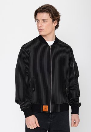 Młody mężczyzna w czarnej bomberce w cienkie prążki z zamkiem błyskawicznym z przodu i kieszenią na suwak na rękawie, patrzy w lewo.