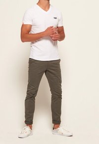 T-shirt blanc à col en V, pantalon slim olive, et baskets blanches avec des accents rouges. Design simple, coupe ajustée et bas de pantalon retroussés.