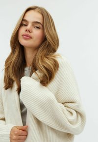 Gilet crémeux chunky avec une texture côtelée, des manches larges et une coupe ample, porté par-dessus un haut gris clair.