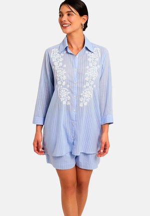 Femme souriante portant une chemise bleu clair à fines rayures avec broderie florale blanche et un short assorti sur un fond uni.