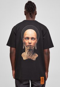 Sort bomulds t-shirt med et stort, detaljeret grafik af et ansigt og tribal-elementer på ryggen. Etiketten med logoet er synlig ved sømmen.