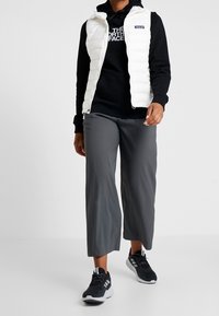 Fleece bianco sopra una felpa nera con cappuccio, abbinato a pantaloni larghi grigi e scarpe sportive nere con dettagli bianchi.