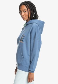 Mėlynas medvilninis džemperis su kengūros kišenėle, puošia spalvingi kalnų grafikai krūtinėje, reguliuojama kapošmens virvutė ir raukšlėti rankogaliai.