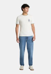 T-shirt bianco a maniche corte con dettagli grigi e logo, abbinato a jeans in denim azzurri e sneakers color beige. Vestibilità casual con forma rilassata.