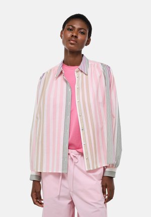 Femme portant un t-shirt rose sous une chemise ample à boutons à rayures multicolores à manches longues et un pantalon rose clair sur un fond uni.